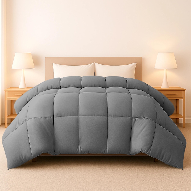Op de afbeelding: Een grijze, gewatteerde dekbedovertrek op een bed, met een houten hoofdbord en twee witte kussens. Twee bijpassende nachtkastjes met lampen flankeren het bed. De dekbedovertrek is effen van kleur en heeft een gestikt, rasterachtig patroon.