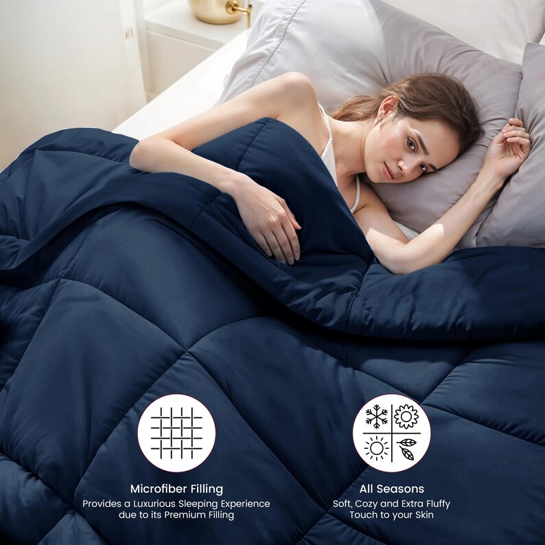 Op de afbeelding: Een marineblauwe quilt met een gewatteerd patroon. Het ligt op een bed met grijze kussens. De tekst op de afbeelding luidt "Microfiber Filling" en "All Seasons".