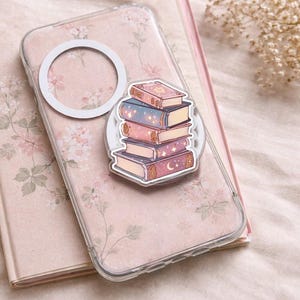 Op de afbeelding: Een doorzichtige telefoonhoes met een bloemenpatroon en een witte ring. Een stapel geïllustreerde boeken met een hemels thema is aan de hoes bevestigd. De boeken hebben roze, blauwe en gouden accenten.
