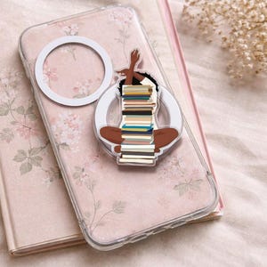 Op de afbeelding: Transparante telefoonhoes met een bloemenpatroon en een witte ringgreep. Een sticker van een persoon die met een stapel boeken zit, is aan de hoes bevestigd. De hoes ligt op een roze bloemennotitieboek.