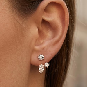 May include: A gold ear jacket earring featuring a round brilliant-cut stud at the top and a dangling component below the earlobe. The lower piece consists of a marquise-cut stone and a smaller round-cut stone set in a delicate gold metal frame.