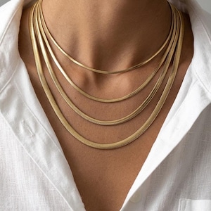 Könnte beinhalten: Ein mehrlagiges Set aus vier goldfarbenen Schlangenkettchen in unterschiedlichen Stärken und Längen, die um den Hals getragen werden. Die Ketten haben ein flaches, ineinandergreifendes Glieddesign, das eine glatte und flexible Oberfläche bildet.