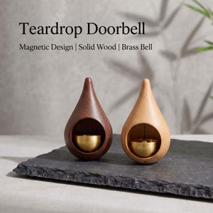 Könnte beinhalten: Zwei tropfenförmige Türklingeln aus massivem Holz, eine dunkelbraun und eine hellbeige, jeweils mit einer polierten Messingglocke in einer kreisförmigen Aussparung. Der Text lautet Teardrop Doorbell, Magnetic Design | Solid Wood | Brass Bell.