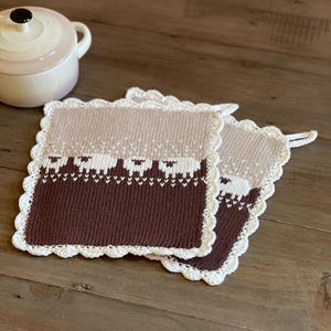 Può includere: Due presine quadrate lavorate a maglia marrone e beige con bordo smerlato bianco. Le presine presentano un disegno di pecora. Una piccola pentola rosa chiaro con manico nero è sullo sfondo.