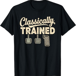 May include: Black t-shirt featuring the text "Classically TRAINED" in a vintage font above an illustration of three car pedals. The cream-colored design contrasts with the black fabric. A humorous design for driving enthusiasts.
