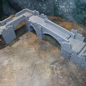 May include: A grey miniature bridge with a castle-like design. The bridge features an arched centre, a ramp, and stairs. The model is made of a light grey material and is designed for tabletop gaming.