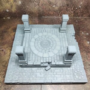Könnte beinhalten: Ein graues, 3D-gedrucktes Miniatur-Tabletop-Gaming-Geländestück. Das Stück hat ein zentrales kreisförmiges Design, vier Säulen und ein strukturiertes, steinartiges Aussehen. Die Basis misst etwa 25 cm x 25 cm.