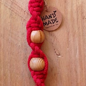 Op de afbeelding: Rode macramé sleutelhanger met twee houten kralen. De sleutelhanger is handgemaakt en heeft een metalen ring aan de bovenkant. Een klein, rond label met de woorden "HAND MADE" en "Especially for You" is bevestigd.