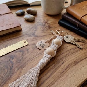 Op de afbeelding: Een handgemaakte macramé sleutelhanger met twee houten kralen en een kwast. De sleutelhanger is bevestigd aan een goudkleurige sluiting met sleutels. Een label met de tekst "Hand Made" is aan de sleutelhanger bevestigd.