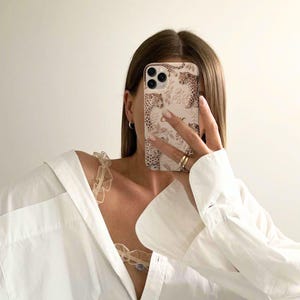 May include: A beige phone case with a leopard print design is held up in front of a person's face. The case features a repeating pattern of leopard spots and floral accents. The person is wearing a white button-down shirt and gold rings.