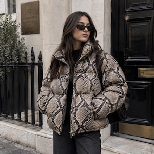 May include: A puffer jacket featuring a tan and black snakeskin print pattern, a high collar, and a front zip closure. The jacket is worn over a black top and paired with dark trousers, complemented by black rectangular sunglasses.