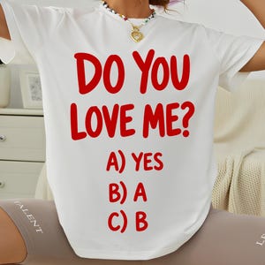 Op de afbeelding: Een wit T-shirt met korte mouwen en opvallende rode tekst met de tekst DO YOU LOVE ME? gevolgd door een meerkeuzelijst: A) YES, B) A en C) B. Het T-shirt heeft een ronde hals en een relaxte pasvorm met een grafisch ontwerp op de voorzijde.