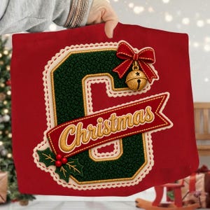 May include: A red fabric item featuring a large, dark green letter C with a textured surface, outlined in cream and gold. A red ribbon bow with a gold jingle bell and a red banner with the gold embroidered text Christmas are attached.