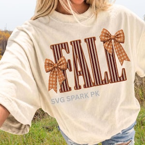 May include: A cream-colored short-sleeve t-shirt featuring the word FALL in large, vertical-striped maroon and white letters. Two orange and white gingham bows are attached to the letters F and L. The text SVG SPARK PK is printed below.