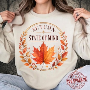 Puede incluir: Una sudadera de cuello redondo color crema con un gráfico central de una gran hoja de arce naranja rodeada por una corona circular de hojas y bayas de otoño. El texto "AUTUMN STATE OF MIND" está impreso en color marrón sobre la hoja.