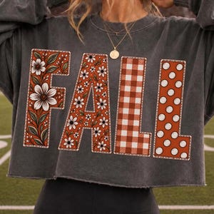 May include: A charcoal gray long-sleeve cropped sweatshirt featuring the word FALL in large block letters. The F has a floral pattern, the A has a daisy print, the L has a brown gingham check, and the second L has a white polka dot pattern.