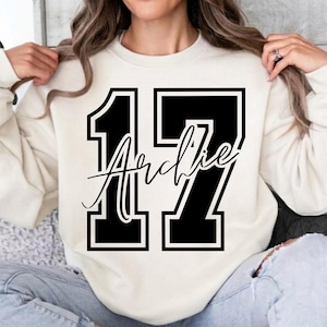 Può includere: Una felpa girocollo color crema con un grande numero 17 in stile varsity stampato in nero sul davanti. Sovrapposto al centro del numero 17, il nome Archie è scritto in un carattere corsivo nero con un contorno bianco.
