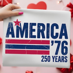 May include: A white t-shirt featuring a distressed navy blue graphic that reads AMERICA, with a red star above the letter A. Below, red and navy blue horizontal stripes sit next to the text '76 and 250 YEARS in navy blue block lettering.