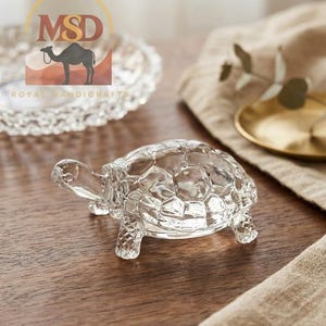 May include: A clear glass turtle figurine featuring a textured shell with a geometric pattern and smooth, defined limbs. The transparent sculpture is crafted with intricate detailing on the head and feet, showcasing a reflective surface.