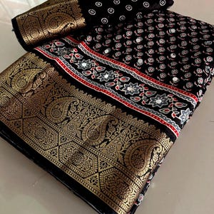 May include: A folded black fabric piece featuring a wide gold metallic border with intricate paisley patterns. The main section displays a repeating floral print in white and red, accented with small circular mirrors throughout the textile surface.