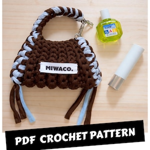 Op de afbeelding: Een bruine en lichtblauwe gehaakte tas met een gevlochten handvat en de tekst "MIWACO." Een klein flesje oogdruppels en een witte tube met een zilveren dop zijn ook zichtbaar. De tekst "PDF CROCHET PATTERN" is ook aanwezig.