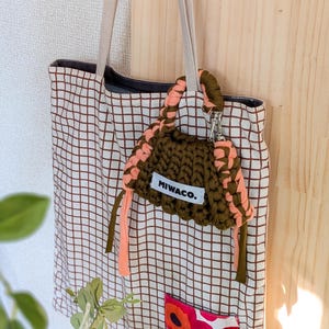 Op de afbeelding: Een tote bag met een bruin rasterpatroon en een kleine, olijfgroene en perzikkleurige gehaakte tas eraan bevestigd. De tas heeft de tekst "MIWACO." op een wit label. De tote bag heeft beige bandjes.