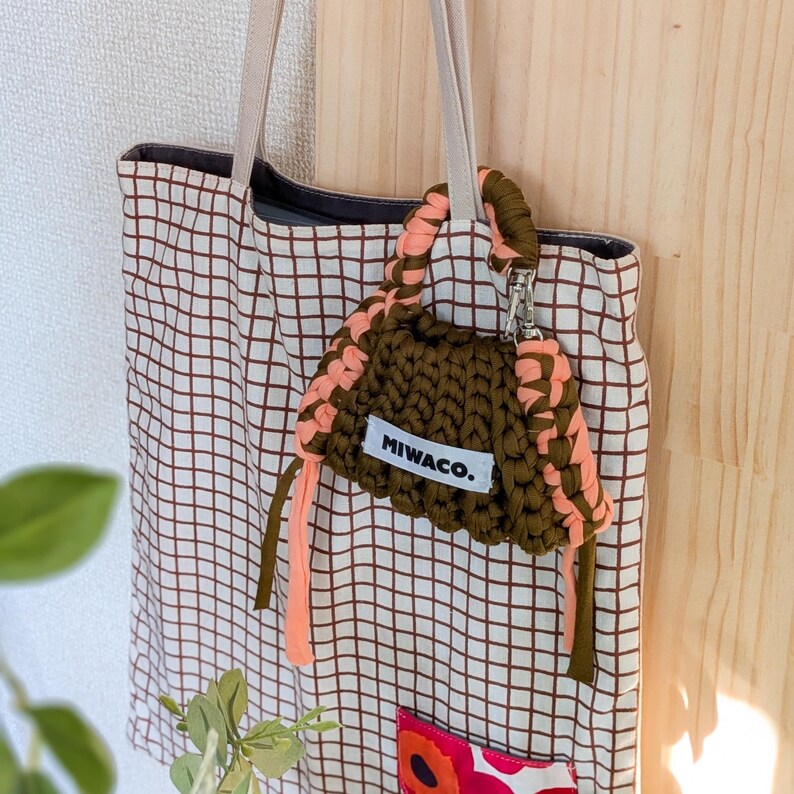 Op de afbeelding: Een tote bag met een bruin rasterpatroon en een kleine, olijfgroene en perzikkleurige gehaakte tas eraan bevestigd. De tas heeft de tekst "MIWACO." op een wit label. De tote bag heeft beige bandjes.