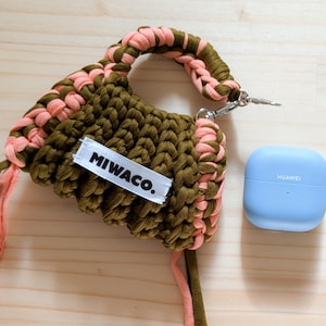 Op de afbeelding: Een handgemaakte macram&eacute; tas in olijfgroen en perzik met een zilveren sluiting en de tekst "MIWACO." Een lichtblauwe Huawei case ligt naast de tas. De tas heeft een gevlochten handvat en een bijpassende riem.