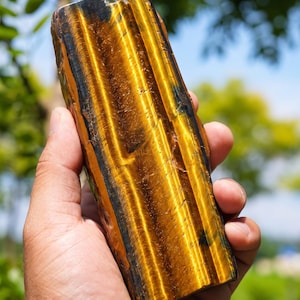 May include: A polished, elongated piece of tiger's eye gemstone featuring prominent golden-yellow and dark brown chatoyant bands that create a shimmering, reflective effect across the smooth, vertical surface of the stone.