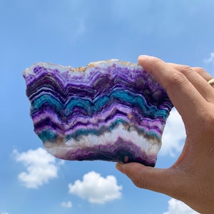 May include: A polished slice of fluorite mineral featuring distinct, wavy bands of deep purple, vibrant teal, and translucent white. The stone displays a natural, jagged top edge and a smooth, flat surface, showcasing intricate, layered color patterns.