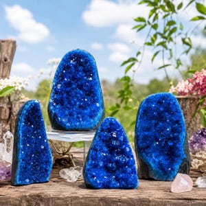 May include: Four vibrant blue geode specimens with crystalline interiors are displayed. The largest geode sits on a clear acrylic stand, while three smaller, pointed geode pieces are arranged around it on a weathered wooden surface.