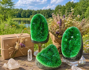 May include: Three vibrant green crystal geodes with sparkling interiors, one mounted on a gold metal stand, another on a clear acrylic base, and a third resting on a wooden surface alongside a brown gift box and small clear quartz points.