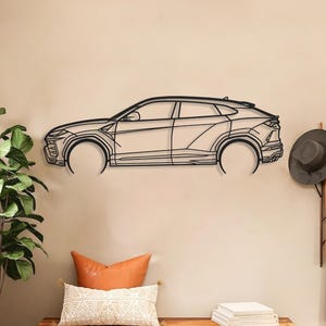 Könnte beinhalten: Ein schwarzes Metall-Wandkunstwerk, das das Seitenprofil eines Autos darstellt. Das Kunstwerk ist an einer beigefarbenen Wand über einer Holzbank mit Kissen und einer Pflanze angebracht. Ein Hut und ein Mantel hängen rechts an der Wand.