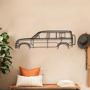May include: A black metal wall sculpture of a car's side profile, mounted on a beige wall. The artwork is above a wooden bench with cushions and baskets. A potted plant and coat rack are also in the scene. The car is a Land Rover.