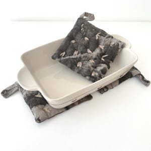 Peut inclure: Un plat de cuisson rectangulaire en céramique blanche accompagné de deux maniques en tissu matelassé. Les maniques présentent un motif moucheté gris foncé et noir avec de petits accents brodés clairs et une boucle de suspension.