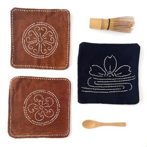 May include: Two square brown fabric coasters with white stitched circular patterns, one square dark blue fabric coaster with a white stitched floral design, a bamboo matcha whisk, and a small wooden spoon arranged on a plain white surface.