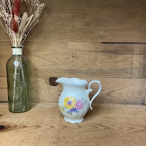 Op de afbeelding: Een witte keramische kan met een bloemenontwerp met gele en paarse bloemen. De kan heeft een geschulpte rand met een blauwe rand en een gebogen handvat. Links staat een groene glazen fles met gedroogde bloemen.