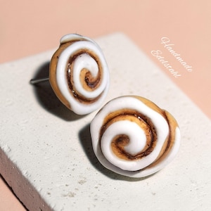 Könnte beinhalten: Ein Paar Ohrstecker in Form von Zimtschnecken mit beigefarbenem Teig, dunkelbraunen Zimtspiralen und weißem Zuckerguss. Der Text "Handmade Edelstahl" ist in der oberen rechten Ecke des Bildes in der Nähe der Ohrringe zu sehen.