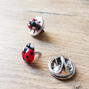 Peut inclure: Deux épinglettes en forme de coccinelle avec un corps rouge à pois noirs et des détails blancs. Une épinglette est vue de côté avec son attache métallique argentée, tandis que l'autre est séparée, montrant la tige et le fermoir argenté.