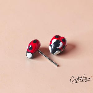 May include: A pair of stud earrings shaped like ladybugs, featuring a glossy red finish with black spots and white accents. Each earring has a silver-toned metal post attached to the back. The text Craft Nitzer is visible in the bottom right corner.