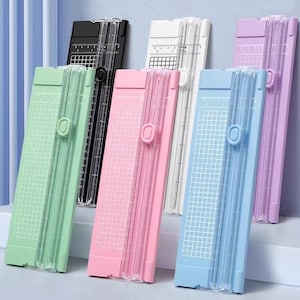 May include: A collection of paper trimmers in various colors, including green, black, white, pink, blue, and purple. Each trimmer has a clear plastic blade guard and a grid for precise cutting. The trimmers are designed for paper and craft projects.