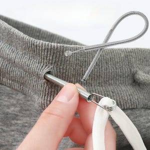 May include: Close-up of a gray sweatpants waistband with a metal drawstring threader inserted. The threader is a coiled spring with a loop at one end and a clasp holding a white drawstring.