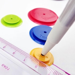 May include: A set of circular plastic spacers in green, red, blue, and yellow, each featuring a central hole and size markings. A white pen tip rests in the center of the yellow 5mm spacer, positioned next to a clear ruler with inch markings.