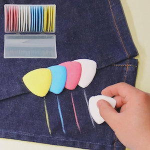 May include: A collection of colorful tailor's chalk in a clear plastic case, with individual pieces in pink, white, blue, and yellow. A hand holds a white chalk piece, demonstrating its use on dark blue fabric.