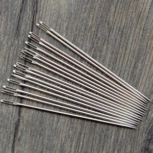 May include: A collection of twelve silver-colored metal sewing needles arranged in a fan pattern. Each needle features a long, slender body with a sharp, pointed tip at one end and an elongated eye opening at the opposite end for threading.