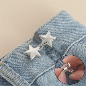 May include: A silver-toned metal waist tightening accessory featuring two connected star-shaped buttons. The fastener is shown attached to the waistband of light blue denim jeans, with an inset detail displaying the back pin mechanism.