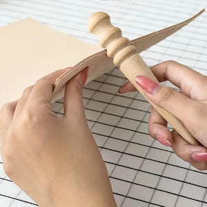 May include: A light-colored wooden leather burnisher tool with a grooved handle and a tapered point, being used to smooth the edge of a thick, natural-toned piece of vegetable-tanned leather. The tool features a rounded, ribbed grip section.