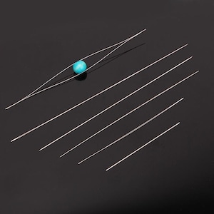 May include: A set of long, thin, silver-colored beading needles arranged diagonally. One needle is threaded through a single, round, bright turquoise bead, while the other five needles are straight and unthreaded, all featuring sharp, pointed tips.