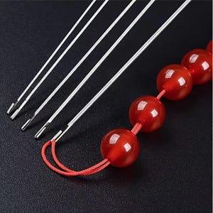 May include: A set of four silver-coloured metal needles with open-eye ends, arranged diagonally. One needle is threaded with a red cord featuring four smooth, round, translucent red beads, creating a continuous loop of vibrant colour and metallic texture.