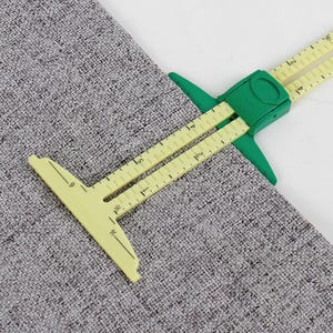 May include: A yellow and green seam gauge tool measuring fabric. The tool has a green sliding component and yellow arms with inch measurements. The fabric is a gray woven material. The tool is designed for accurate seam allowances.